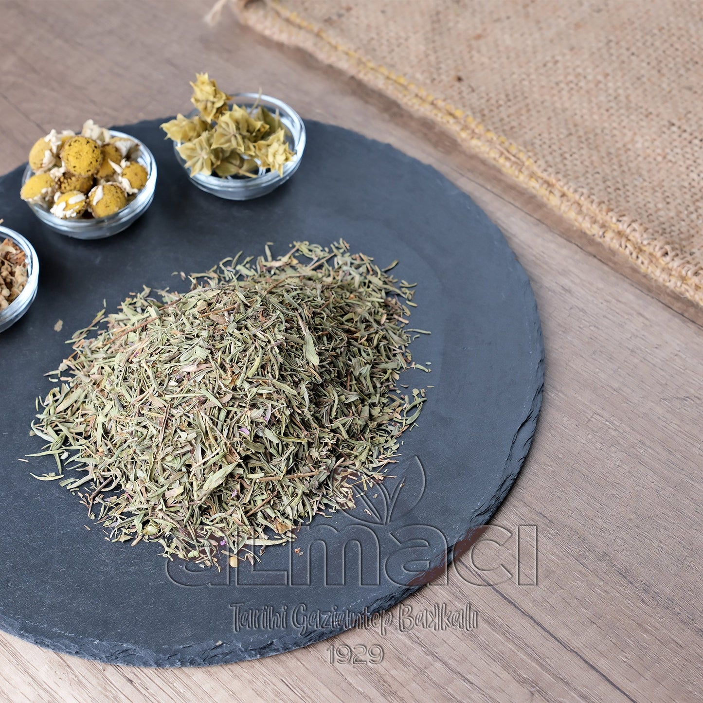 Yuvarlak siyah bir taş plaka üzerinde biberiyeler yığın halinde duruyor. Yanında cam kaselerde çeşitli kurutulmuş bitkiler var. Arka planda hasır kumaş parçası bulunuyor.