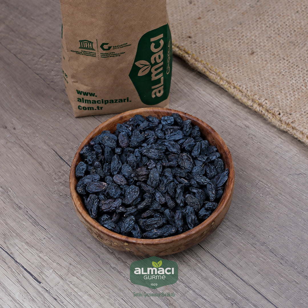 Tahta bir kasede siyah çekirdeksiz kuru özbek üzümler, ahşap zemin üzerinde duruyor. Yanında, "www.almacipazari.com.tr" yazılı kahverengi bir kağıt paket var. Paketin yanında hasır dokulu bir kumaş yer alıyor.