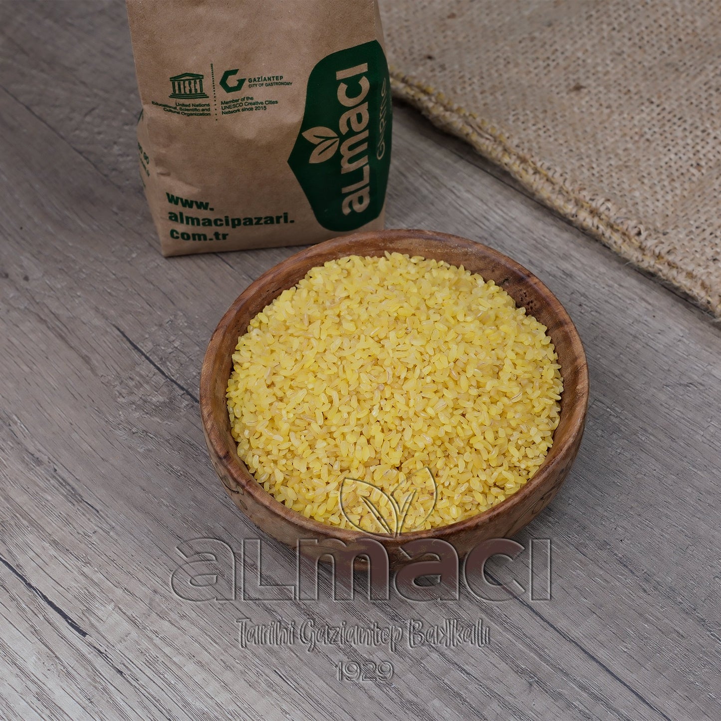 Tahta bir kasede iri taneli sarı altın bulgur taneleri duruyor. Yanında, üzerinde "Almacı", "www.almacipazari.com.tr", "Gaziantep" yazılı kahverengi bir paket var. Arka planda hasır bir kumaş bulunuyor.