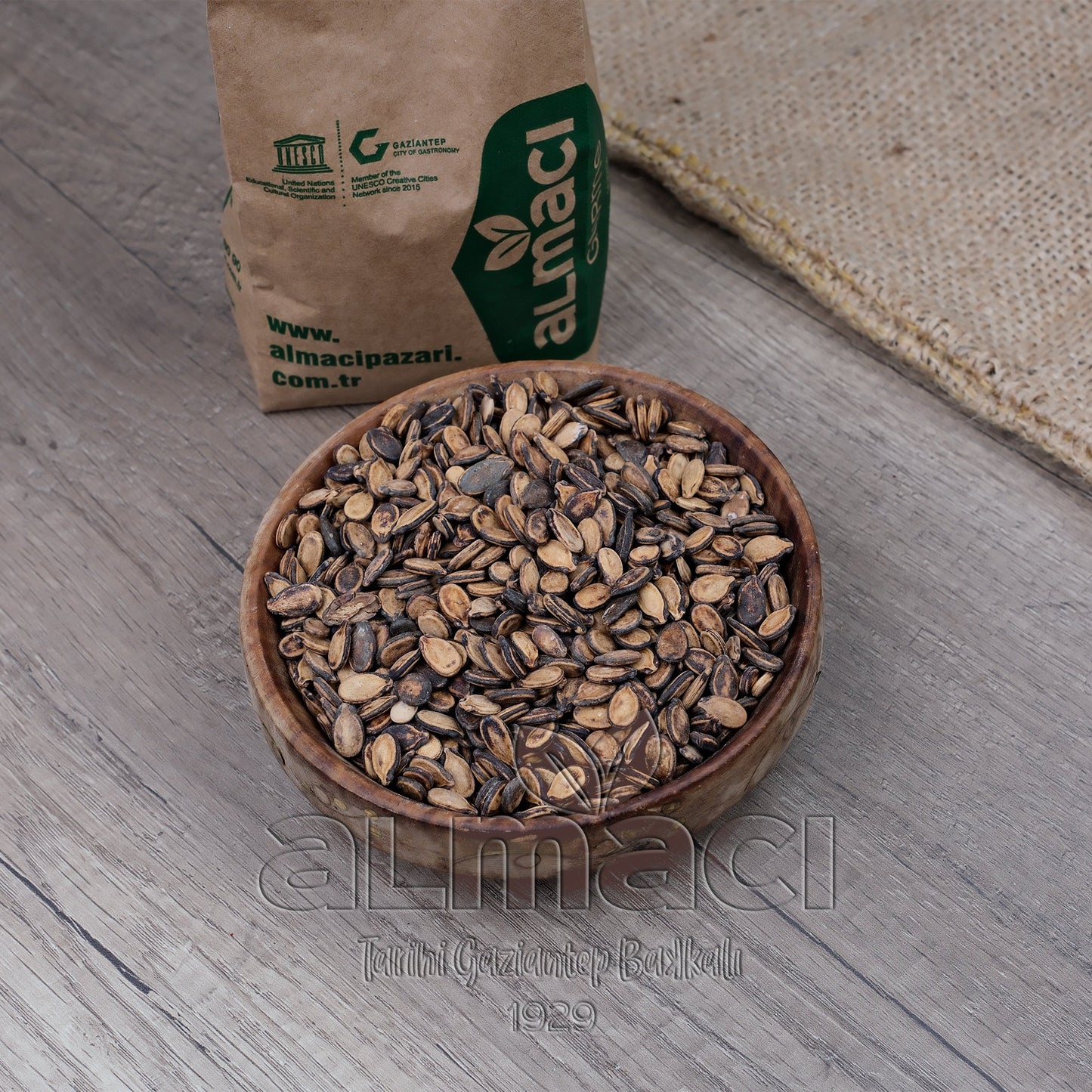 Ahşap bir kasede kavrulmuş karpuz çekirdeği bulunuyor. Zeminde bir kilim ve ahşap zemin görülüyor.