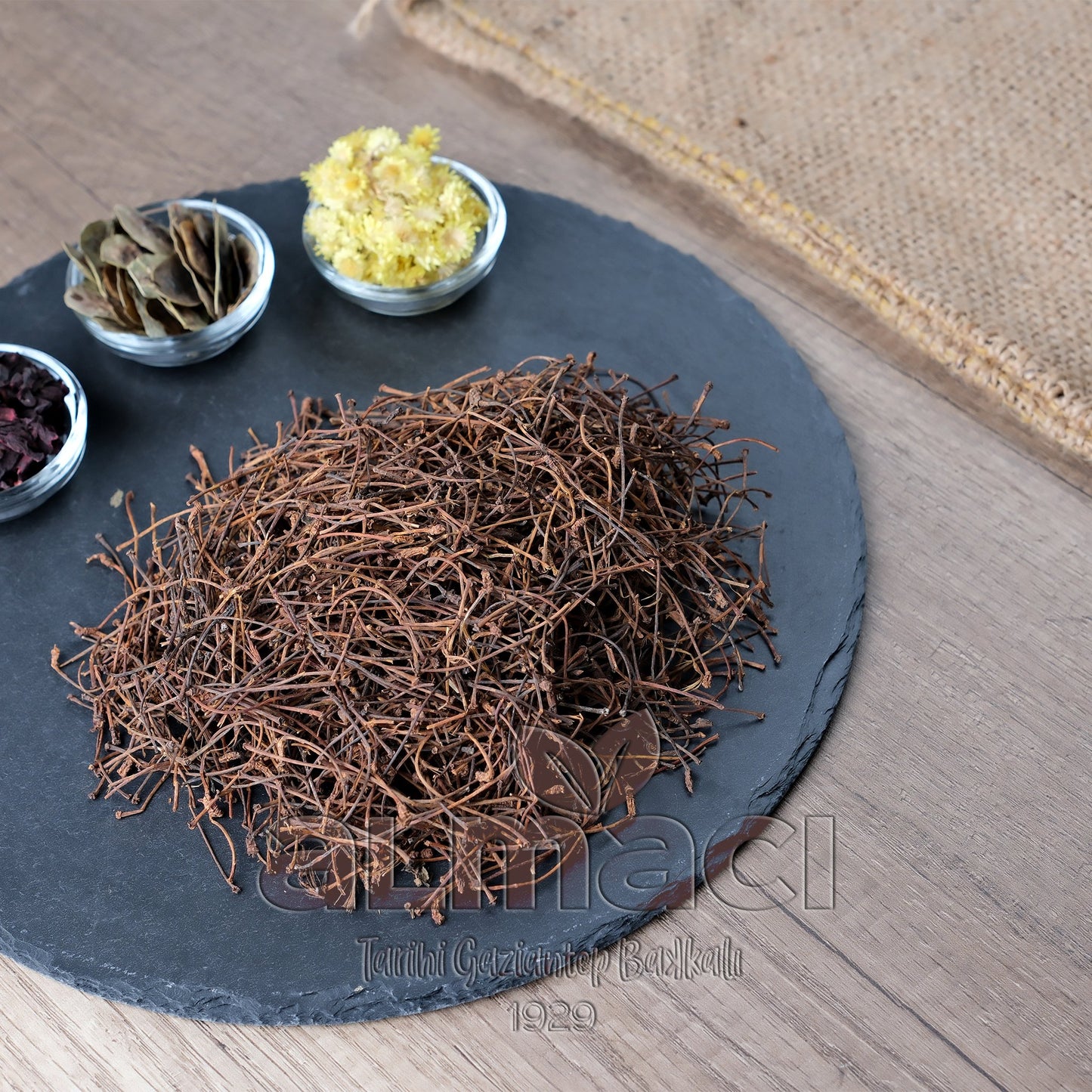 Siyah bir tabak üzerinde kurutulmuş kiraz sapı yığılmış, yanında küçük kaselerde çeşitli bitkiler var; arka planda hasır kumaş.