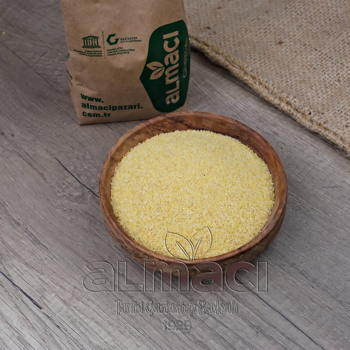 Tahta kasede duran köftelik bulgur, bir masa üzerinde sergileniyor. Yanında, üzerinde “almacipazari.com.tr” yazılı bir kağıt torba bulunuyor.