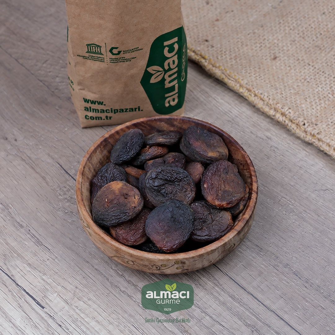 Tahta kasede kuru kayısı günkurusular durmakta. Yanında kahverengi kağıt paket var. Zemin ahşap olup doğal bir ortamda.