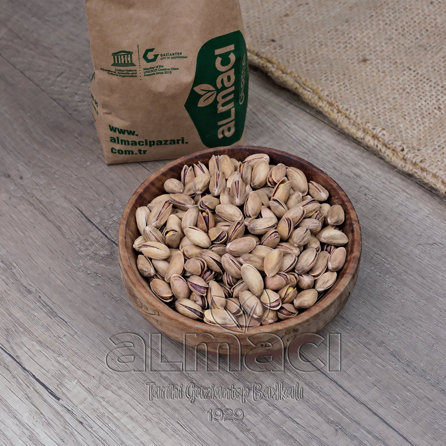 Ahşap bir kasede kabuklu özel Antep fıstığı duruyor. Zeminde ahşap parkeler ve dokulu bir kumaş görülüyor.