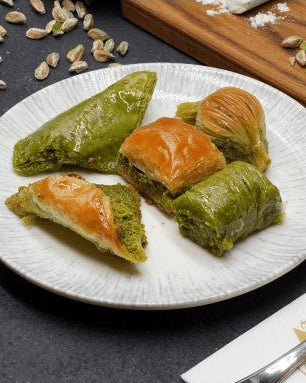 Tabakta yeşil fıstıklı özel baklava çeşitleri duruyor. Yanında kesme tahtasında fıstıklar ve toz fıstık var. Çatal-bıçak ve peçete masanın köşesinde.