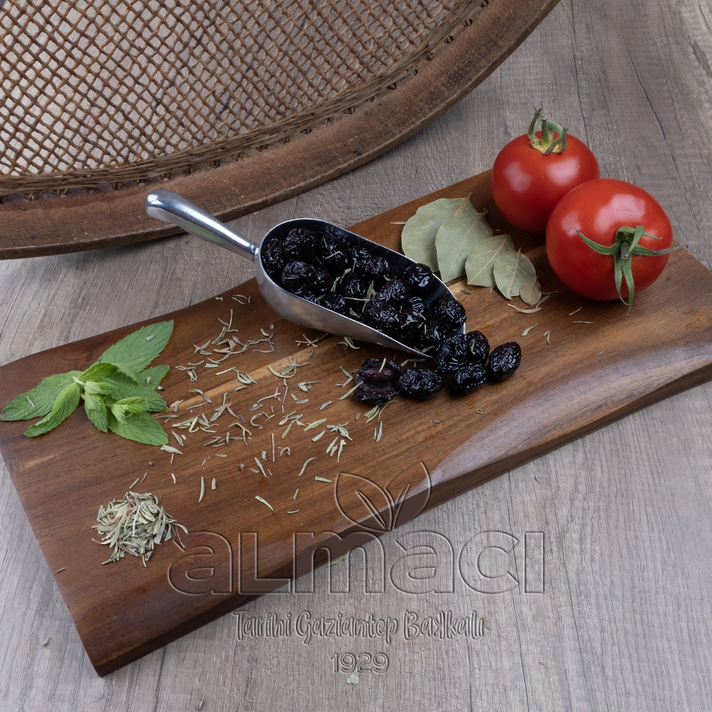 Metal bir kepçede köy selesi siyah zeytinler, yanında domates, defne yaprağı ve nane, ahşap bir tahta üzerinde yer alıyor.