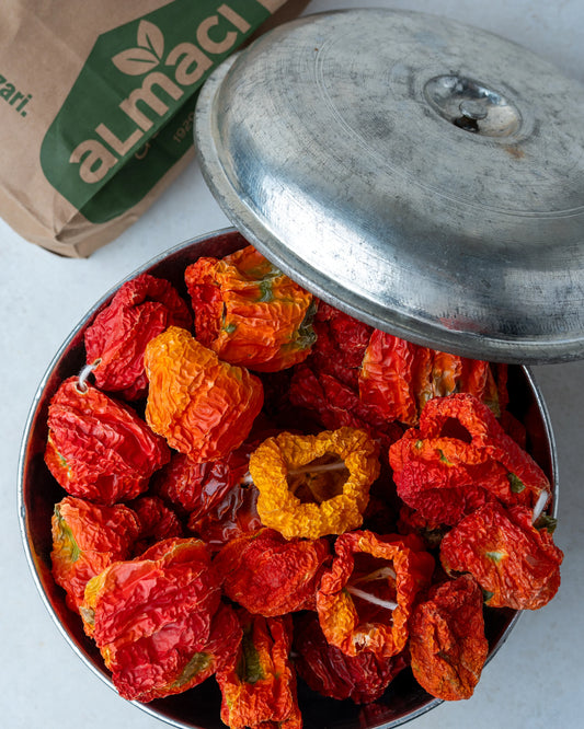 Metal kasede sunulan kırmızı kuru biberler ve arka planda Almacı Pazarı ambalajı, güneşte kurutulmuş ince kabuklu acı dolmalık sebze.