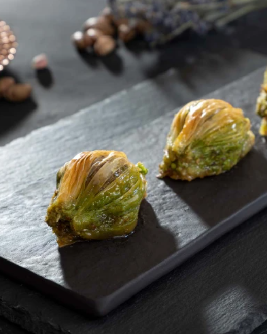 Taş bir tabak üzerinde iki adet fıstıklı midye baklava dilimi duruyor. Arkada fındıklar ve bakır bir tabakta dekoratif nesneler bulunuyor, loş bir ortamda çekilmiş.