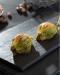 Taş bir tabak üzerinde iki adet fıstıklı midye baklava dilimi duruyor. Arkada fındıklar ve bakır bir tabakta dekoratif nesneler bulunuyor, loş bir ortamda çekilmiş.