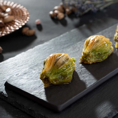 Taş bir tabak üzerinde iki adet fıstıklı midye baklava dilimi duruyor. Arkada fındıklar ve bakır bir tabakta dekoratif nesneler bulunuyor, loş bir ortamda çekilmiş.