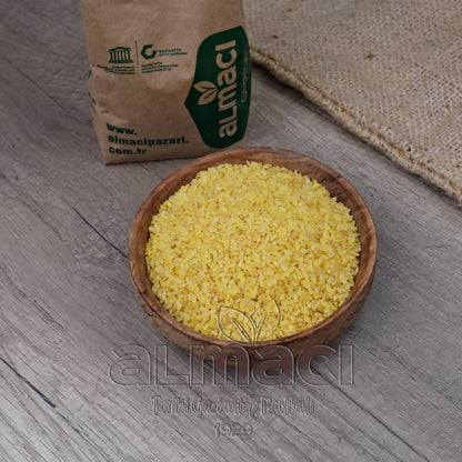 Tahta bir kasede iri taneli sarı altın bulgur taneleri duruyor. Yanında, üzerinde "Almacı", "www.almacipazari.com.tr", "Gaziantep" yazılı kahverengi bir paket var. Arka planda hasır bir kumaş bulunuyor.