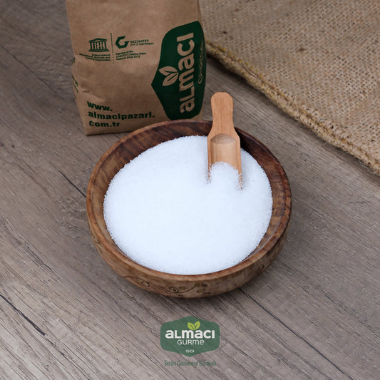 Tahta kapta limon tuzu, üzerinde küçük tahta bir kaşık bulunmaktadır. Yanında "almacipazari.com.tr" yazılı kahverengi kağıt paket ve hasır malzeme yer almakta. 