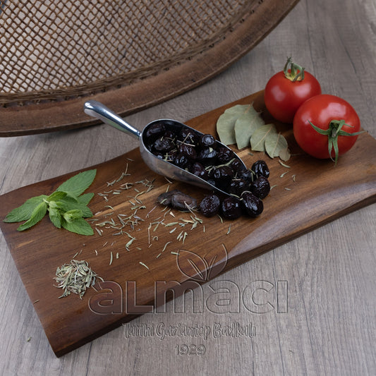 Metal kepçe içindeki siyah baskı xl zeytinler, tahta kesme tahtasında defne yaprakları, taze nane dalları, ve iki domatesle çevrili. 